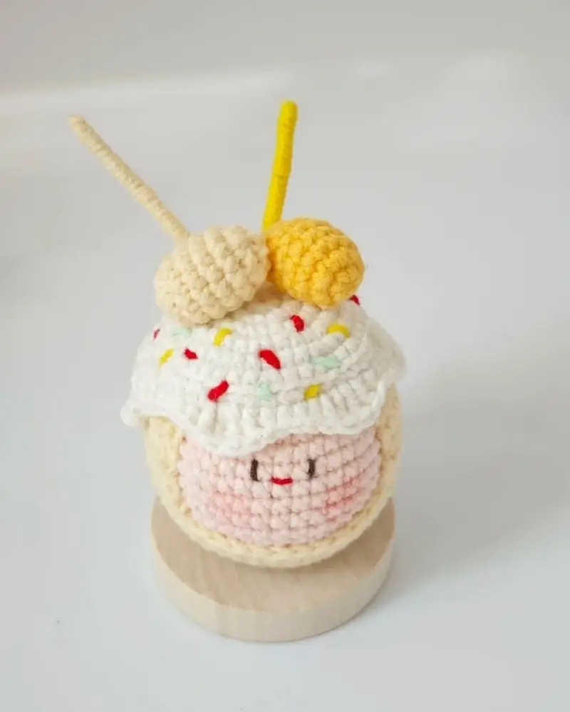 Crocheted cupcake toy, pink frosting, sprinkles, yellow cherries.