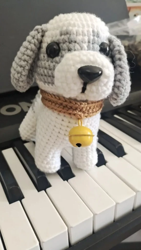 Crocheted dog toy, gray and white, with brown collar and yellow bell, sitting on piano keys.