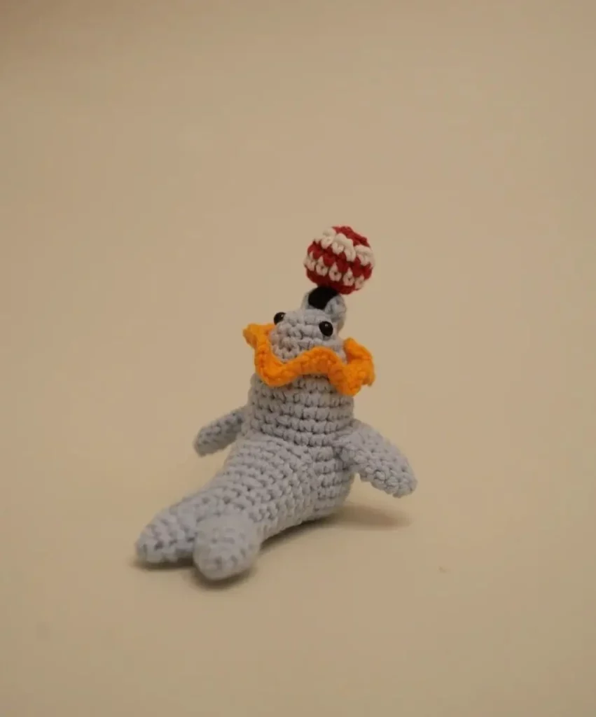 Crocheted seal puppet, juggling a red ball, light gray with orange trim.