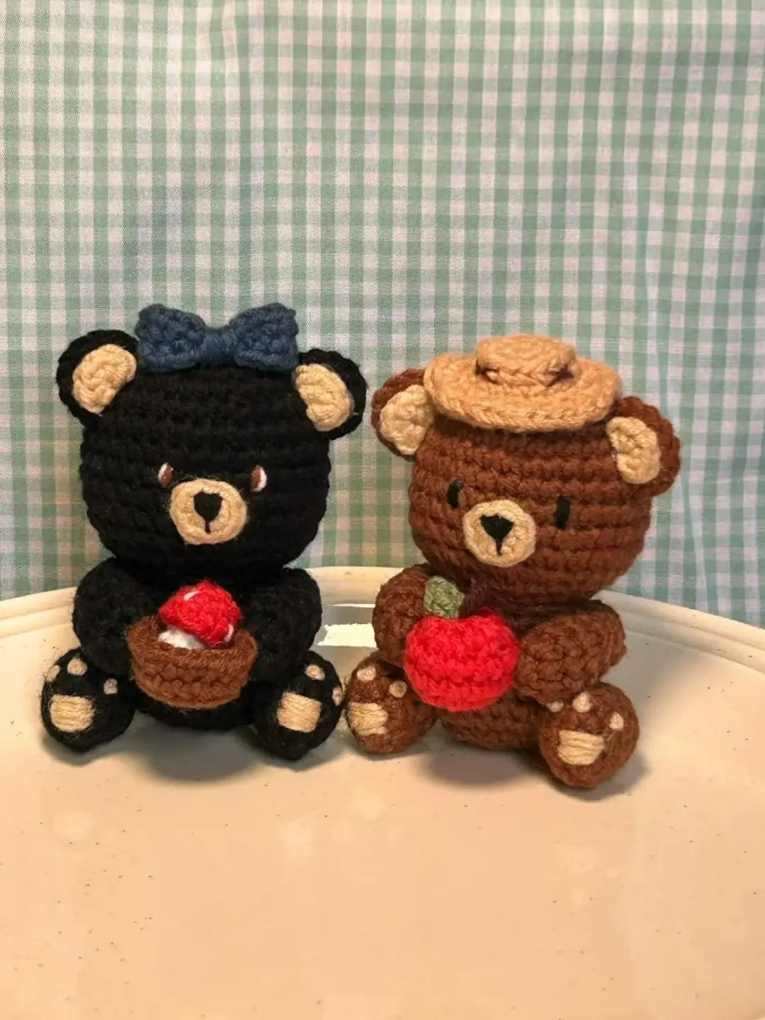 Two crocheted teddy bears, one black with a blue bow, the other brown with a straw hat, holding apples.