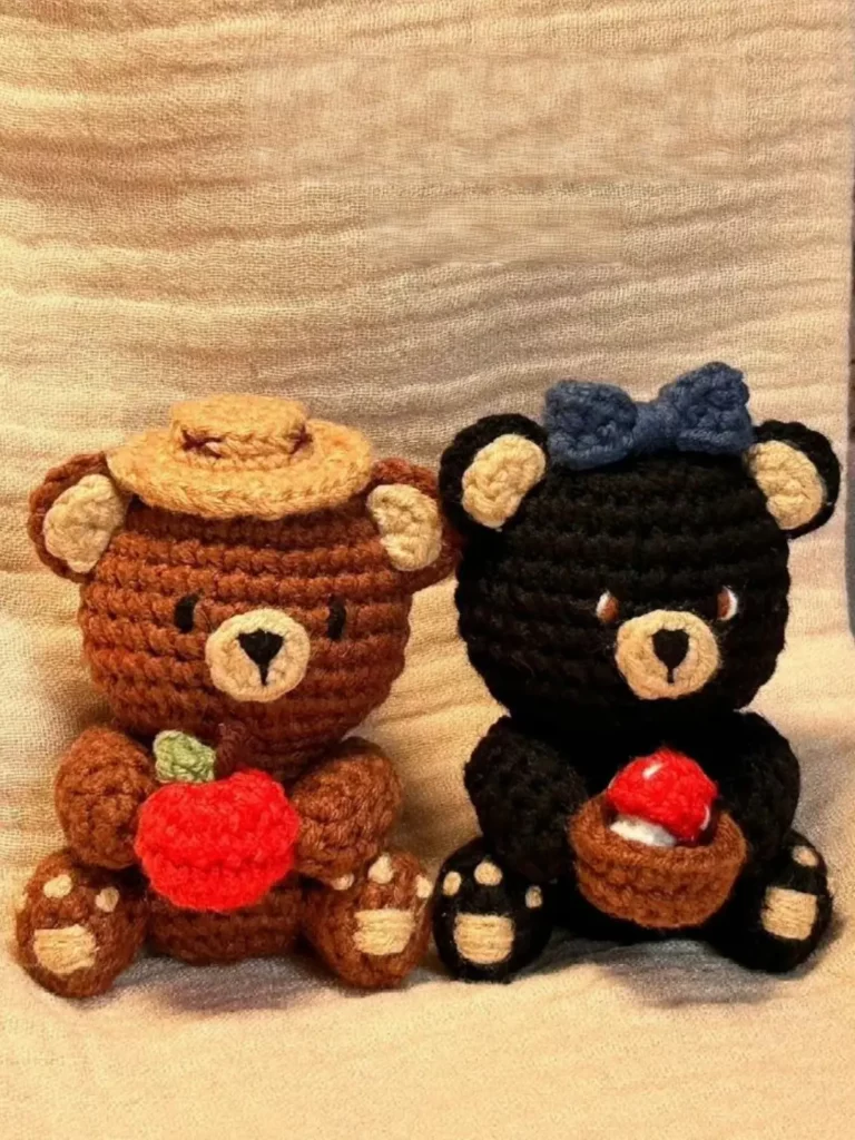 Two crocheted teddy bears, one brown with a hat and an apple, the other black with a blue bow and a mushroom.