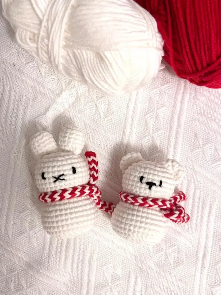 Two crocheted animals, a rabbit and a bear, with red and white scarves, on a white knitted blanket.  Winter animals.