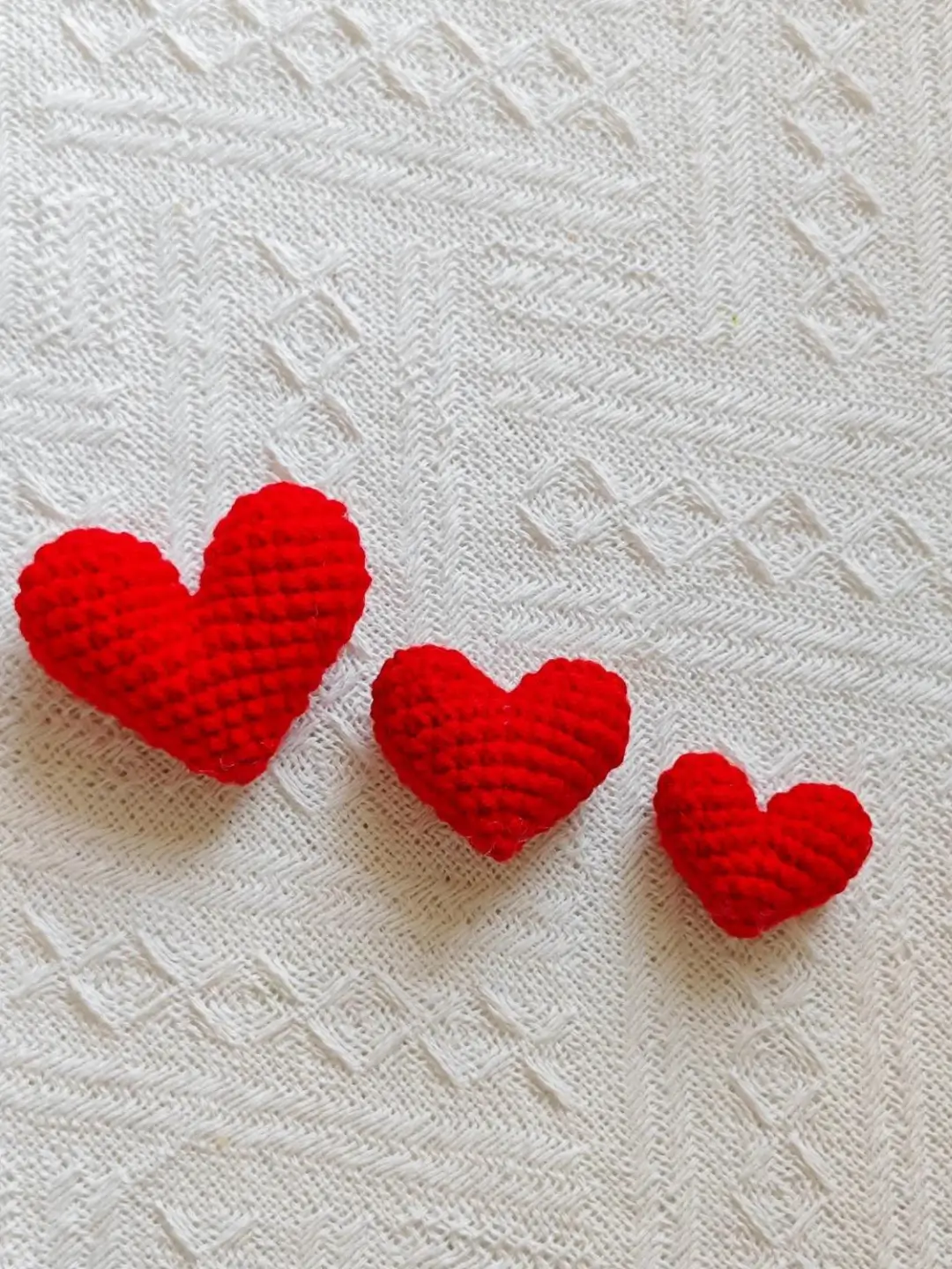 Three red crocheted hearts on white knitted fabric.